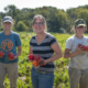 three people standing in a farm field holding handfuls of red bell peppers