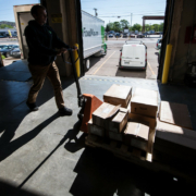 Person moving boxes with a pallet jack on a loading dock with trucks in the background