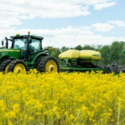 tractor pulls a no-till drill through a field of blooming yellow mustard below blue sky with clouds