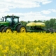 tractor pulls a no-till drill through a field of blooming yellow mustard below blue sky with clouds