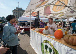 man standing in open booth tent with different kinds of squash on a table in front of him and audience members facing him.