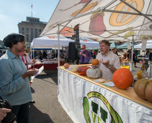 man standing in open booth tent with different kinds of squash on a table in front of him and audience members facing him.