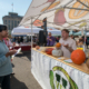 man standing in open booth tent with different kinds of squash on a table in front of him and audience members facing him.