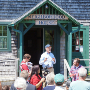 A speaker addresses a crowd standing outdoors in front of a wood-shingled building