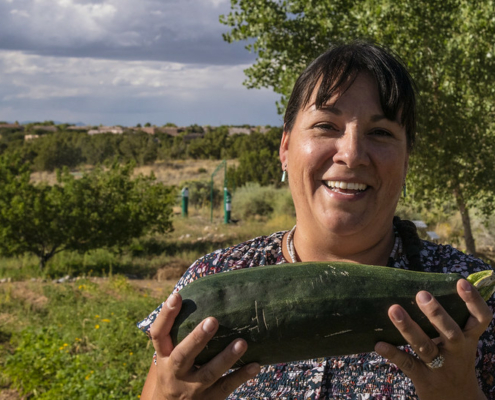 smiling person holding a zucchini in foreground, with plants, bushes, and sky behind.