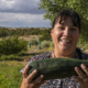 smiling person holding a zucchini in foreground, with plants, bushes, and sky behind.