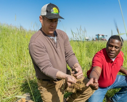 Two men crouch in a green field with blue sky above, holding a handful of soil that they are looking at.