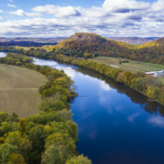 High-angle view looking down on a river with trees along the banks, farm fields, and hills, under a blue sky with clouds.