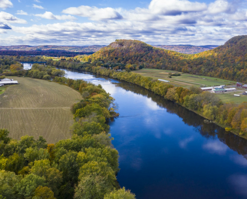 High-angle view looking down on a river with trees along the banks, farm fields, and hills, under a blue sky with clouds.