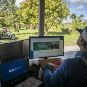 A man with his back to the camera sits in front of computer screens with trees and farm equipment in the background