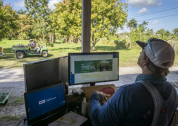 A man with his back to the camera sits in front of computer screens with trees and farm equipment in the background