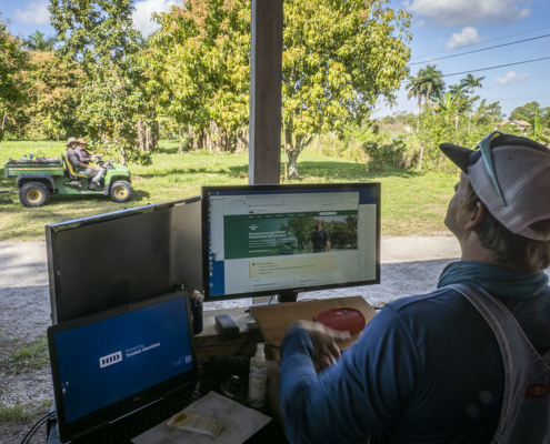 A man with his back to the camera sits in front of computer screens with trees and farm equipment in the background