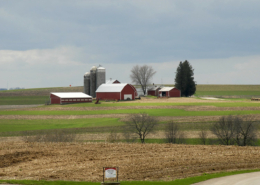 distant view of farmstead with bars, silos, and trees, with blue sky in background and fields in foreground.