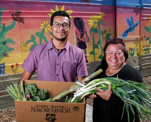 two people holding a cardboard box of produce pose in front of a colorful mural