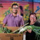 two people holding a cardboard box of produce pose in front of a colorful mural