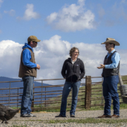 three people stand talking in front of blue sky with white clouds. A duck is in the foreground.