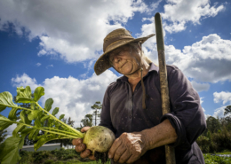 woman holding a radish and a hand tool, against blue sky with white clouds