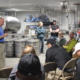 People in chairs listen to a standing speaker in a maple-syrup processing facility