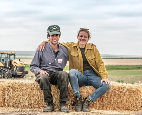 two people with arms around each other's shoulders sit on a bale in front of sky with clouds