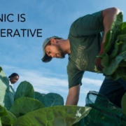 man leaning over green plants against blue sky background with words "Organic Is Regenerative"