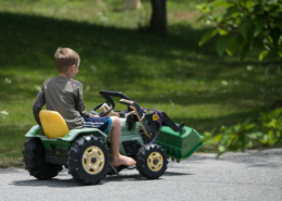 A child riding a toy tractor outdoors on pavement with green grass in background.