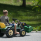 A child riding a toy tractor outdoors on pavement with green grass in background.