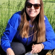 person with long hair, blue shirt, and sunglasses kneeling to plant seeds in the ground