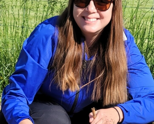 person with long hair, blue shirt, and sunglasses kneeling to plant seeds in the ground