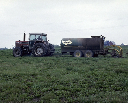 A tractor pulls a tank sprayer across a field, under cloudy sky.