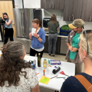 A group of people gathered in a commercial kitchen facility, around a table with ingredients and equipment