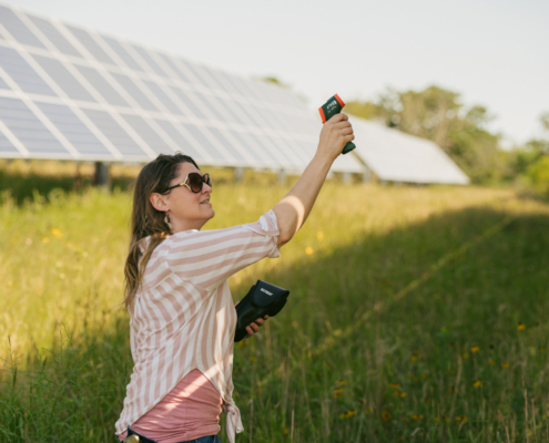 person standing in a field between solar panels, aiming a digital thermometer upward