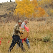 two people walk through a field with a reel of orange flagging, in front of brown vegetation and trees with yellow leaves.