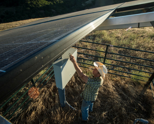 looking down on a person in a cowboy hat making adjustments to controls for a solar panel surrounded by fence.