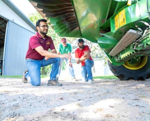 three people look at a rotating mill mounted on the bottom of a combine