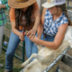 A sheep reclines in a net cradle as two people in hats lean over it to trim hooves.