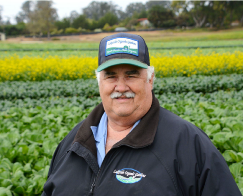 A person in a hat stands in front of a field of vegetables in a head-and-shoulders view.
