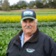 A person in a hat stands in front of a field of vegetables in a head-and-shoulders view.