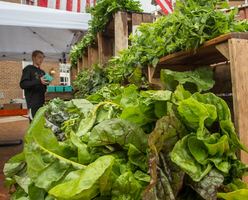 greens displayed at a farmers market with a person and American flags in the background