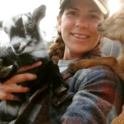 Photo of Danielle holding a black and white goat, with a tan goat leaning on her shoulder.