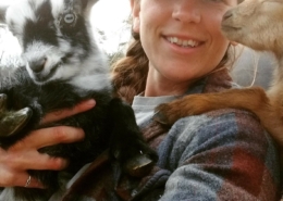 Photo of Danielle holding a black and white goat, with a tan goat leaning on her shoulder.