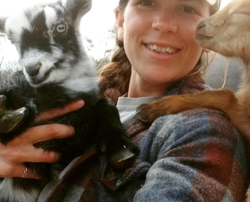 Photo of Danielle holding a black and white goat, with a tan goat leaning on her shoulder.