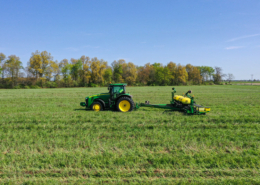 A tractor pulls seeding equipment across a green field under blue sky, with trees on horizon line