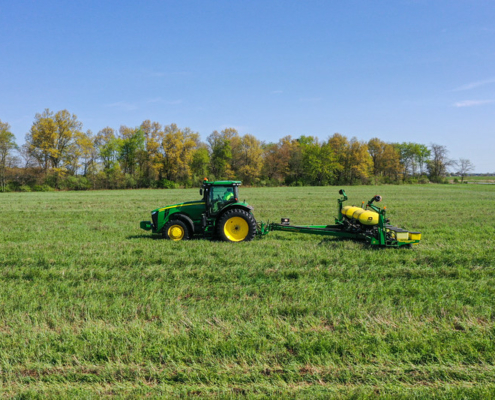 A tractor pulls seeding equipment across a green field under blue sky, with trees on horizon line