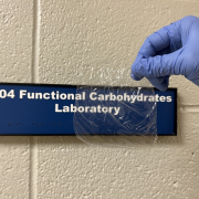 A gloved hand holds a piece of clear plastic in front of a sign on a block wall.