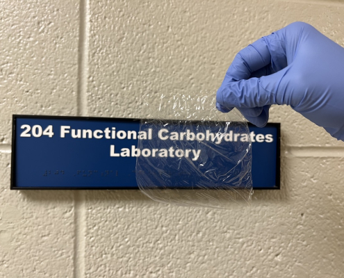 A gloved hand holds a piece of clear plastic in front of a sign on a block wall.