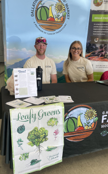 A man and woman sit behind a farmers market table with a Leafy Greens poster displayed. 