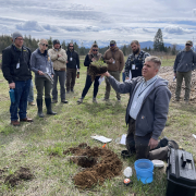 kneeling instructor holds up block of sod to show students, outdoors in field