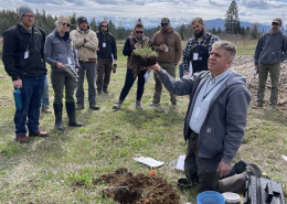 kneeling instructor holds up block of sod to show students, outdoors in field