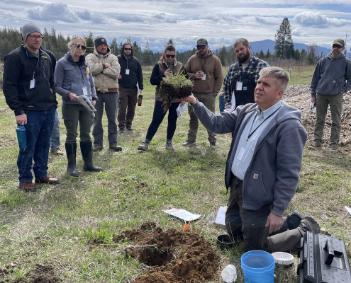 kneeling instructor holds up block of sod to show students, outdoors in field