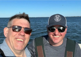Mike and Ray, owners of the oyster farm, in sunglasses, standing on a boat smiling into the camera.